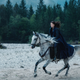 Moiraine rides swiftly on a white horse over a rocky beach, her blue cape flapping in the wind.